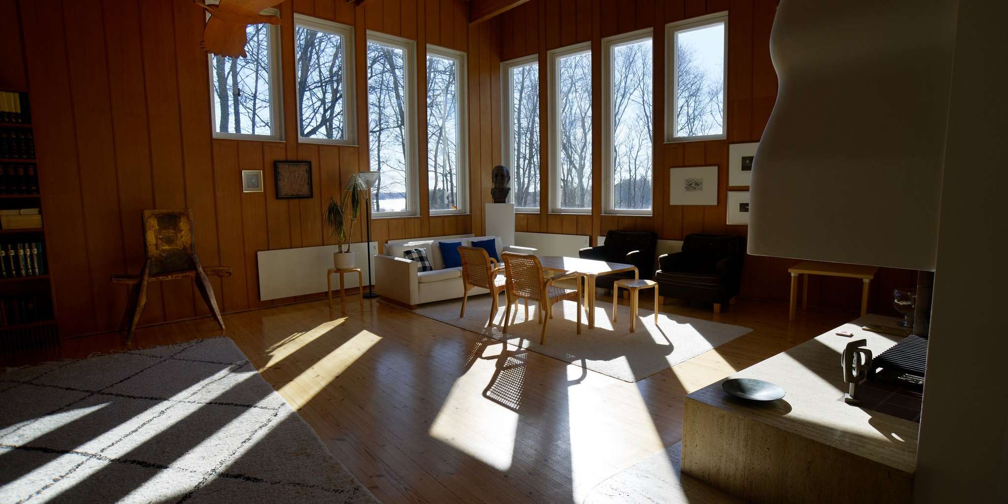 Villa Kokkosen olohuone, jossa suuri ikkuna-alue tuo runsaasti luonnonvaloa. Huoneessa on vaaleita huonekaluja, puisia yksityiskohtia ja moderni takka oikealla.