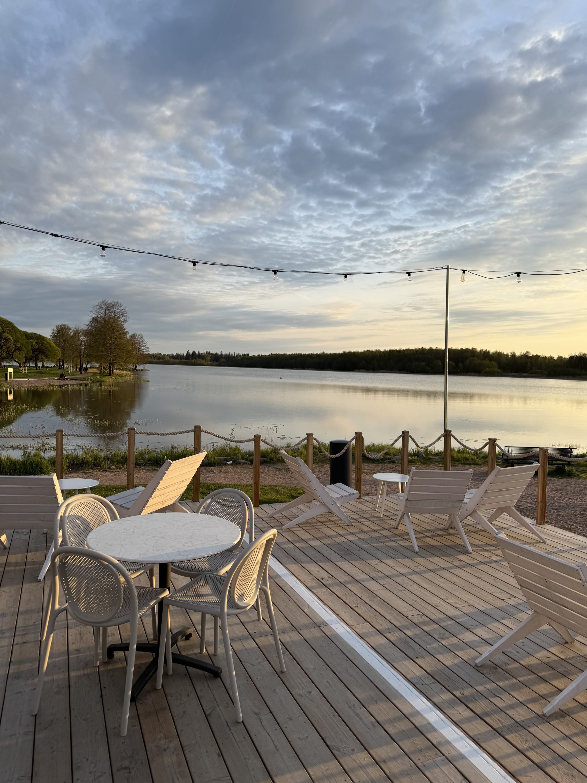 Ilta-auringossa kylpevä puuterassi, jolla on valkoisia tuoleja ja pöytiä. Taustalla rauhallinen järvimaisema ja keväisen vihreä rantaviiva. Taivaalla on ohuen pilviverhon läpi siivilöityvää valoa ja koristevaloja kulkee terassin yllä.