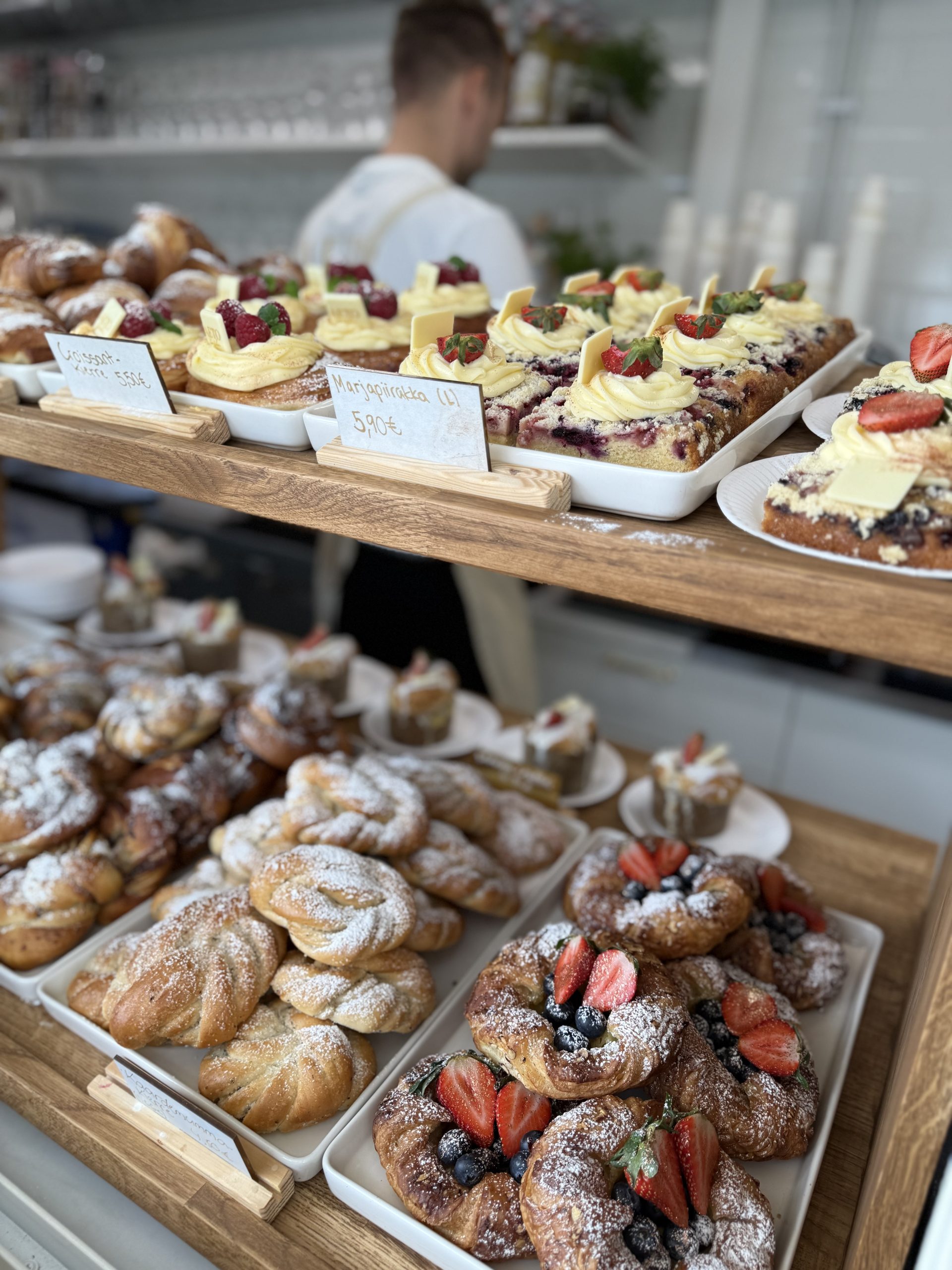 Hyllyllä on runsaasti tuoreita leivonnaisia kahvilassa. Etualalla on marjoilla koristeltuja leivoksia, pullia, croissanteja ja piirakoita. Taustalla näkyy myyjä valkoisessa esiliinassa.