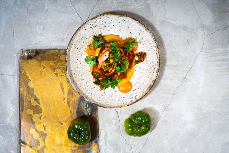 Moderni ravintola-annos keraamisella lautasella, jonka päällä on yrttejä, oranssia kastiketta ja paistettua kanaa tai kalaa. Taustalla on rosoinen betonipinta ja vihreä tomaatti koristeena.