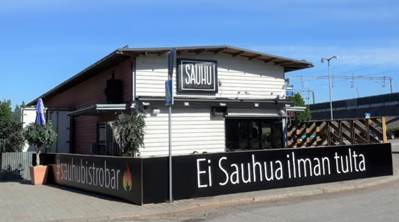 Vaalea puurakennus, jonka seinässä lukee ”SAUHU”. Etualalla musta terassiaitaus, jossa lukee ”Ei Sauhua ilman tulta” ja #sauhubistrobar. Taustalla näkyy junarata ja sinitaivasta.