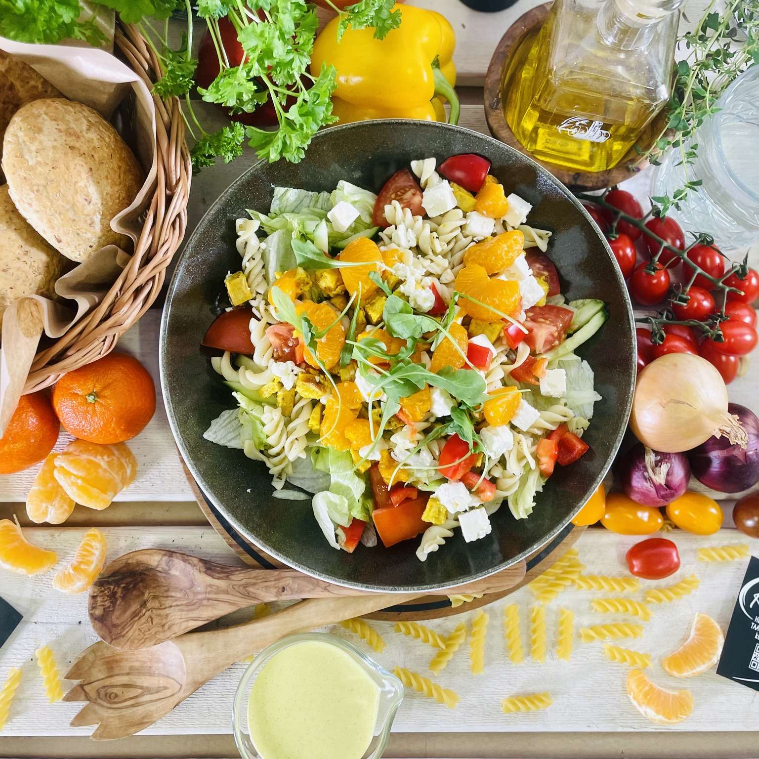 Runsas pastasalaatti kulhossa, jonka ympärillä tuoreita vihanneksia, hedelmiä ja leipää. Kuvassa myös puukauhat, juomapullo ja vihreitä yrttejä.