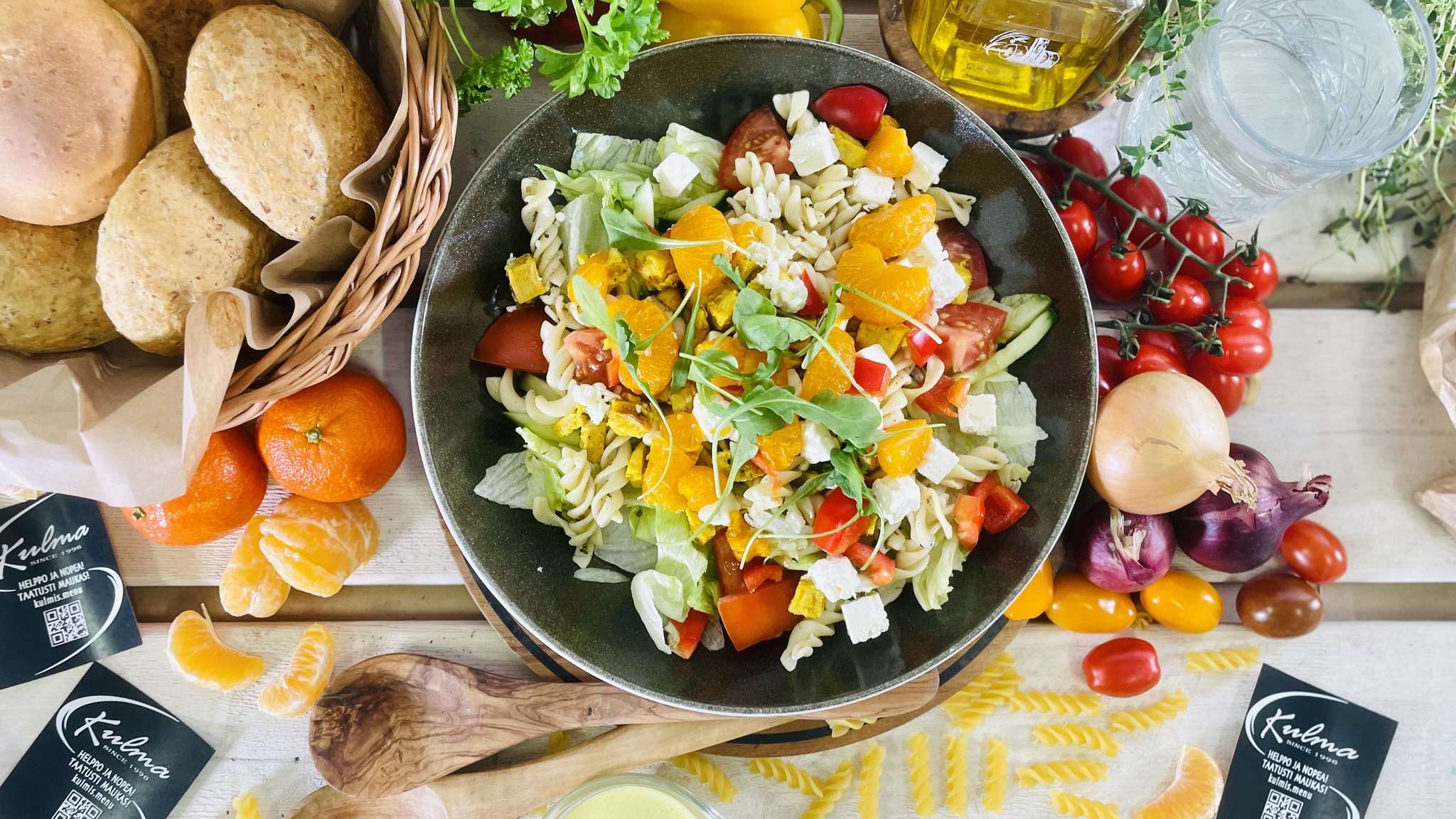 Runsas pastasalaatti kulhossa, jonka ympärillä tuoreita vihanneksia, hedelmiä ja leipää. Kuvassa myös puukauhat, juomapullo ja vihreitä yrttejä.