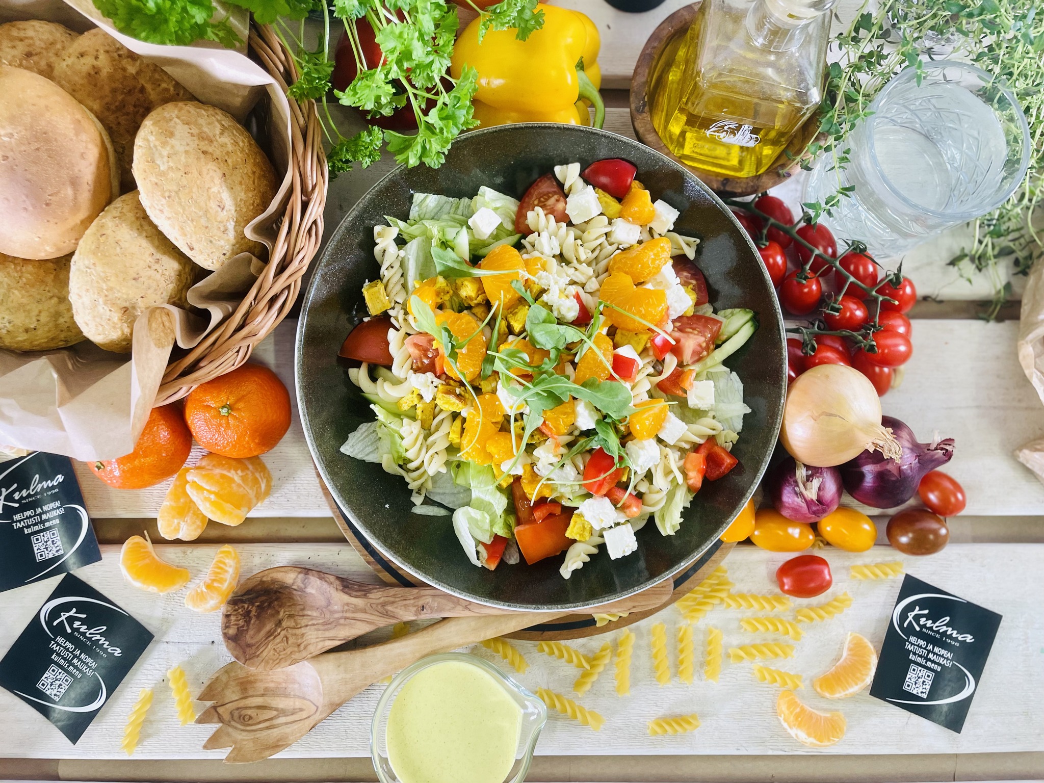 Runsas pastasalaatti kulhossa, jonka ympärillä tuoreita vihanneksia, hedelmiä ja leipää. Kuvassa myös puukauhat, juomapullo ja vihreitä yrttejä.