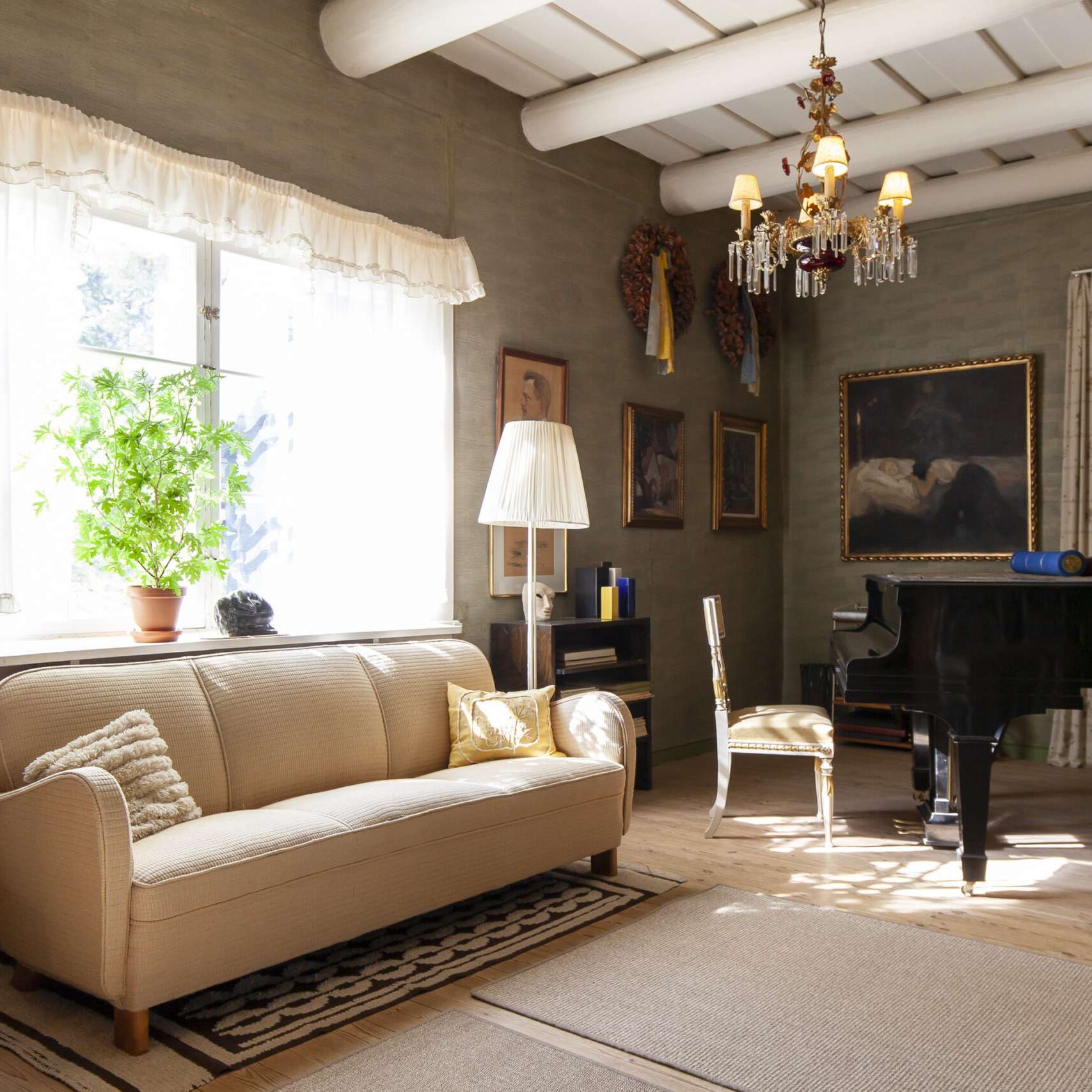 Valoisa sali Ainolan kodissa: ikkunan edess&auml; vaalea sohva ja viherkasvi, seinill&auml; tauluja ja kattokruunu. Oikealla musta flyygeli ja tuoli, sein&auml;ll&auml; suuri maalaus.