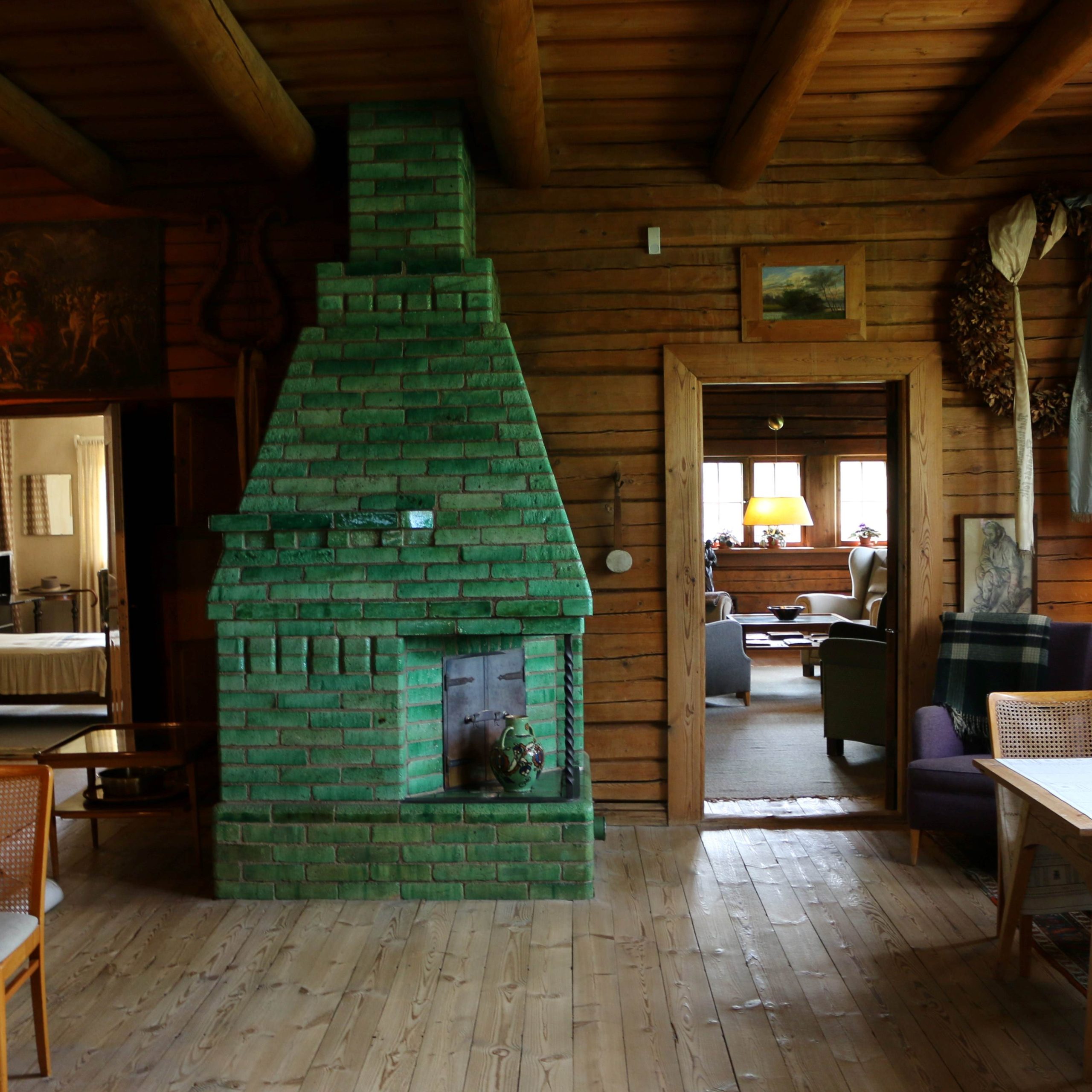 Hirsisein&auml;inen huone, jossa keskell&auml; suuri vihre&auml; kaakeliuuni. Taustalla oviaukko toiseen oleskelutilaan, jossa nojatuoleja ja p&ouml;yt&auml;. Lattia ja katto puuta.
