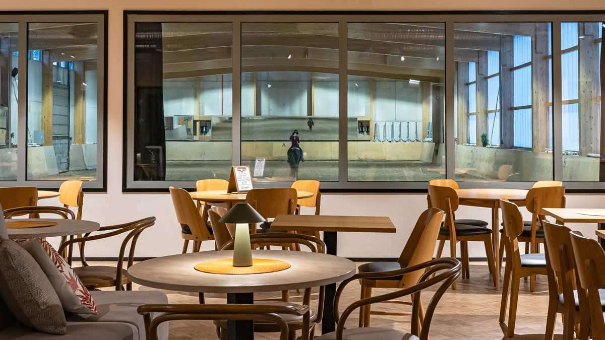 Viihtyis&auml; kahvilatila, jossa on puisia p&ouml;yti&auml; ja tuoleja. Suurista ikkunoista n&auml;kyy ratsastushalli, jossa ratsastaja harjoittelee hevosella.