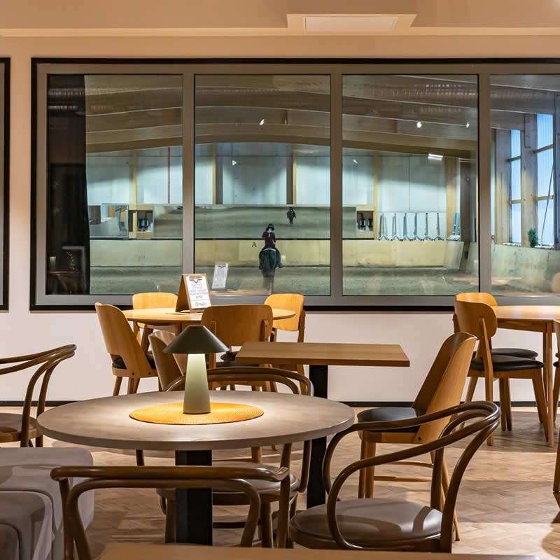 Viihtyis&auml; kahvilatila, jossa on puisia p&ouml;yti&auml; ja tuoleja. Suurista ikkunoista n&auml;kyy ratsastushalli, jossa ratsastaja harjoittelee hevosella.