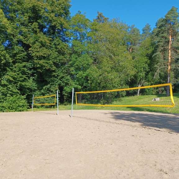 Hiekkainen rantalentopallokentt&auml;, jossa kaksi verkkoa vierekk&auml;in. Taustalla vehre&auml; mets&auml; ja kirkas sininen taivas aurinkoisena p&auml;iv&auml;n&auml;.