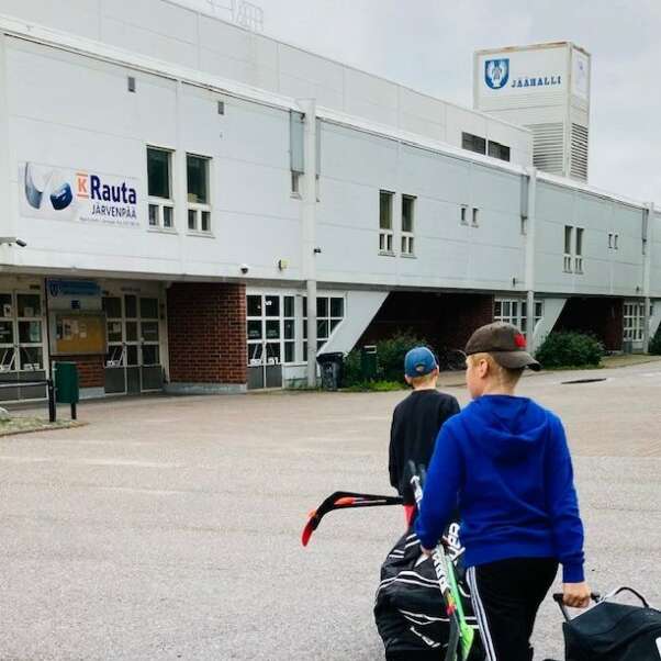 Kaksi lasta k&auml;velee j&auml;&auml;kiekkomailojen ja varustekassien kanssa j&auml;&auml;hallin edustalla.
